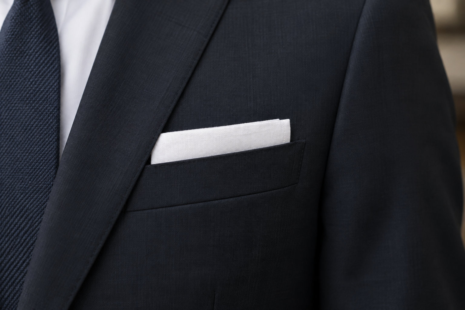 Gros plan sur la poche d’un costume sombre contenant une pochette blanche, avec une chemise blanche et une cravate à motifs.